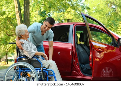 Young Man Helping Disabled Senior Woman In Wheelchair To Get Into Car Outdoors