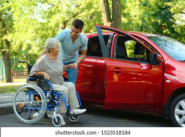 Young Man Helping Disabled Senior Woman In Wheelchair To Get Into Car Outdoors