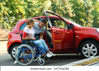 Young Man Helping Disabled Woman In Wheelchair To Get Into Car Outdoors