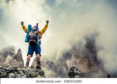 Young man hiker standing on stone mountain with raised up arms with cloudy sky and fog. Yellow jacket, backpack, black beard and beanie. Traveling Dolomites, Italy. - Powered by Shutterstock