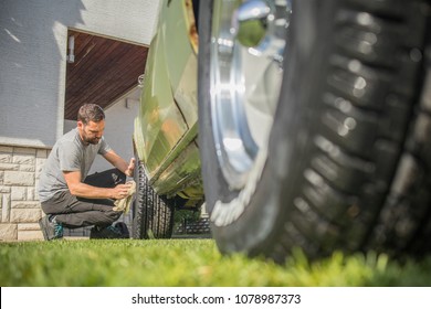 Young Man With A Hip Beard Cleaning A Wheel Of An Old Green Vintage Car On A Home Lawn. Cleaning Muscle Car Tires At Home With A Cloth.