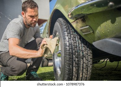 Young Man With A Hip Beard Cleaning A Wheel Of An Old Green Vintage Car On A Home Lawn. Cleaning Muscle Car Tires At Home With A Cloth.