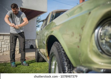 Young Man With A Hip Beard Cleaning A Wringing A Cloth While Cleaning An Old Green Vintage Car On A Home Lawn. Cleaning Muscle Car At Home With A Cloth.