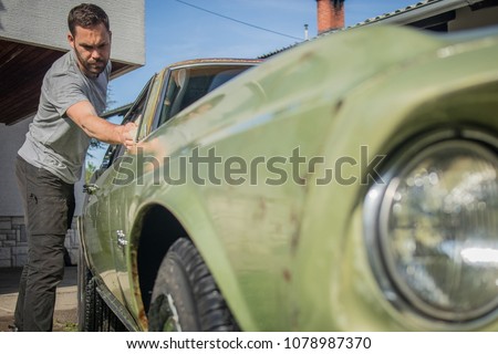 Young man with a hip beard cleaning a window of an old green vintage car on a home lawn. Cleaning muscle car windows at home with a cloth.