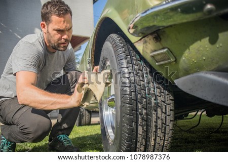 Young man with a hip beard cleaning a wheel of an old green vintage car on a home lawn. Cleaning muscle car tires at home with a cloth.
