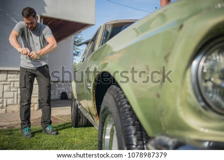 Young man with a hip beard cleaning a wringing a cloth while cleaning an old green vintage car on a home lawn. Cleaning muscle car at home with a cloth.