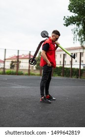 Young Man Hipster Posing With An Electric Scooter. Guy In Trendy Red-black Denim Clothes In Sunglasses In Sneakers With A Scooter On Shoulder Is Resting Standing On A Basketball Court In The City.