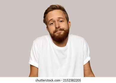 Young Man With Hipster Trendy Haircut And Bearded Chin Shrugging Over Gray Wall At Studio, Pursing Lips, Having No Words To Explain His Behavior, Looking At Camera With Uncertain Smile