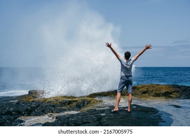 A young man with his arms outstretched standing with his back on the rocks next to an exploding water geyser against the background of the ocean. - Shutterstock ID 2167005975
