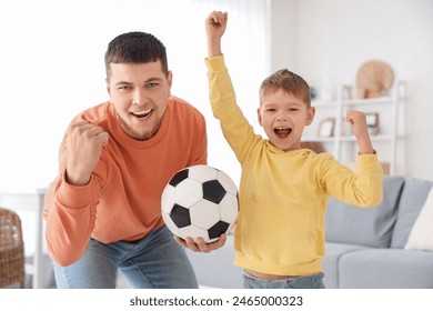 Young man and his little son cheering for football team with soccer ball in living room - Powered by Shutterstock