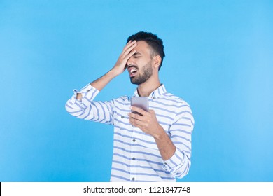 Young Man Hold His Phone Disappointed After Lose The Game Putting Hand On Forehead Sad On A Blue Background.