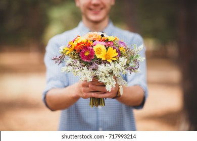 A Young Man Is Holding A Beautiful Bouquet Of Autumn Flowers In His Hand. The Guy Decided To Make A Surprise To His Girlfriend. Romantic Gift For The Girl. Coniferous Forest In Autumn. Background Blur