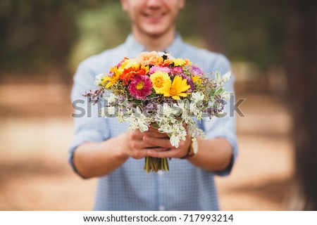 A young man is holding a beautiful bouquet of autumn flowers in his hand. The guy decided to make a surprise to his girlfriend. Romantic gift for the girl. Coniferous forest in autumn. background blur
