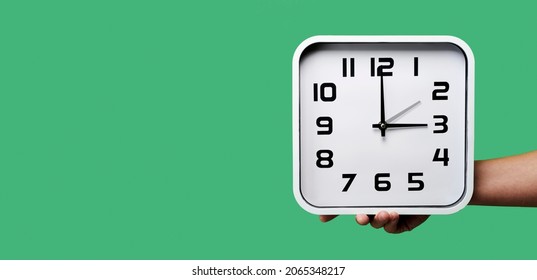 a young man holding a clock being set backward or forward, at the end or beginning of the summer time, on a green background, in a panoramic format to use as web banner or header