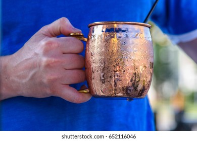 Young Man Holding A Cold Refreshing Moscow Mule In A Bright Shiny Copper Mug Outdoor In Sunlight - Soft Focus