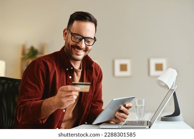 Young man holding credit card and tablet at home at table - Powered by Shutterstock