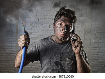 young man holding electrical cable smoking after domestic accident with dirty burnt funny face expression calling desperate with mobile phone asking for help in electricity DIY wrong repairs concept 

