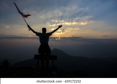 Young Man Holding Flag To Conquer The Mountain. Silhouette Style.