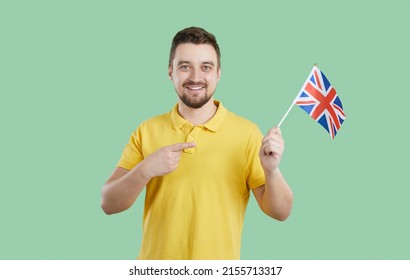 Young Man Holding Flag Of UK. Happy British Guy Smiling And Pointing At Union Jack In His Hand. Studio Portrait Of Student Learning English Language Or Teacher Offering Fun Courses With Native Speaker