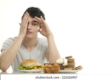 Young Man Holding In Front Lots Of Cookies And A Big Hamburger. Choosing Between Chocolate, Cupcakes, Biscuits And A Burger. Trying To Get Fat Eating Fast Food And Lots Of Sugar