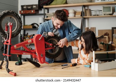 Young man holding hand of his little daughter while checking wheels of her bicycle or repairing it by wooden table in garage - Powered by Shutterstock