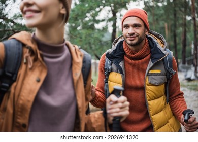 Young Man Holding Hiking Stick And Looking At Happy Girlfriend On Blurred Foreground