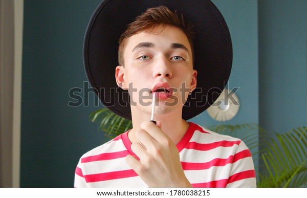 Young\
man holding lipstick or lip gloss and paints lips prepare getting\
ready looking in camera like mirror. Gay make\
up.