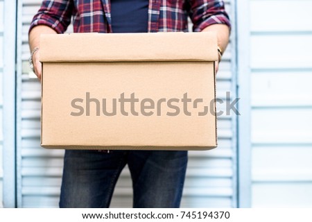 Young man holding a moving cardboard box in front of a storage door.Life style, storage, moving, storing, organizing concept. Space to write