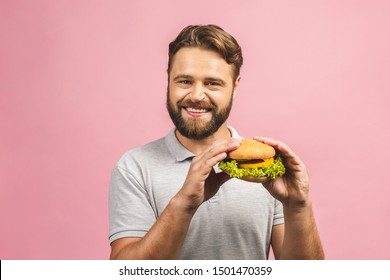 Young man holding a piece of hamburger. Bearded gyu eats fast food. Burger is not helpful food. Very hungry guy. Diet concept. Isolated over pink background. 
