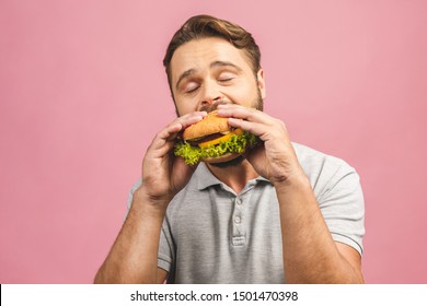 Young man holding a piece of hamburger. Bearded gyu eats fast food. Burger is not helpful food. Very hungry guy. Diet concept. Isolated over pink background.