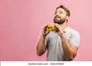 Young Man Holding A Piece Of Hamburger. Bearded Gyu Eats Fast Food. Burger Is Not Helpful Food. Very Hungry Guy. Diet Concept. Isolated Over Pink Background. 