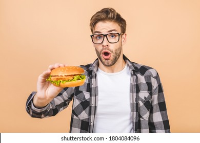 Young Man Holding A Piece Of Hamburger. Student Eats Fast Food. Burger Is Not Helpful Food. Very Hungry Guy. Diet Concept. Isolated Over Beige Background.