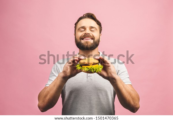 ハンバーガーを持つ若い男性 髭のある牛はファストフードを食べる ハンバーガーは役に立たない食べ物だ お腹が空いた男 国会のコンセプト ピンクの背景に の写真素材 今すぐ編集