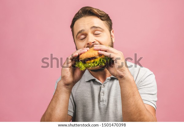 ハンバーガーを持つ若い男性 髭のある牛はファストフードを食べる ハンバーガーは役に立たない食べ物だ お腹が空いた男 国会のコンセプト ピンクの背景に の写真素材 今すぐ編集