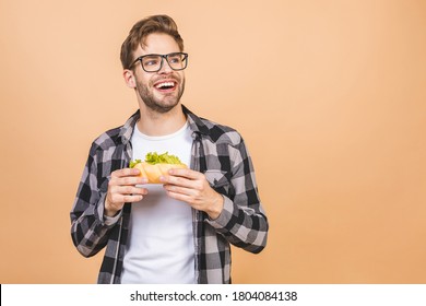 Young man holding a piece of sandwich. Student eats fast food. Burger is not helpful food. Very hungry guy. Diet concept. Isolated over beige background.