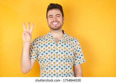 Young Man Holding Pineapple Wearing Hawaiian Shirt Standing Over Isolated Yellow Background Showing And Pointing Up With Fingers Number Three While Smiling Confident And Happy.