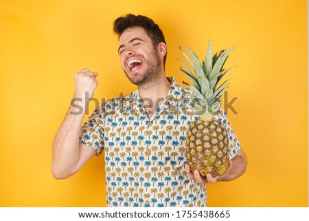 Young man holding pineapple wearing hawaiian shirt standing over isolated yellow background celebrating surprised and amazed for success with arms raised and eyes closed. Winner concept.