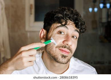 Young man holding a razor while shaving his face, performing a morning grooming routine