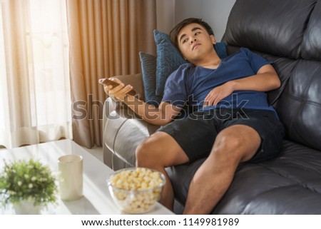 young man holding remote control and watching TV while sitting on sofa in the living room 