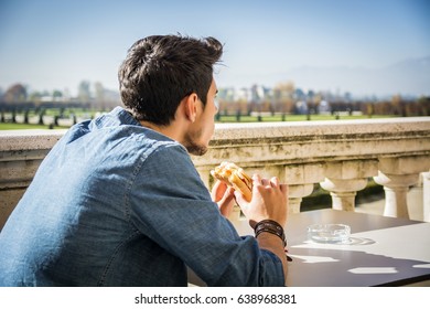 Young man holding sandwich while sitting in cafe terrace, looking to a side seriously. Summertime - Powered by Shutterstock