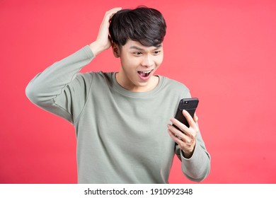 Young man holding smartphone looking at it with a surprised expression, isolated on red background.
