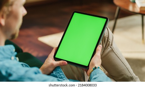 Young Man Holding Tablet Computer with Green Screen Mock Up Display. Male Relaxing at Home, Watching Videos and Reading Social Media Posts on Mobile Device. Close Up Static Over the Shoulder Footage. - Powered by Shutterstock