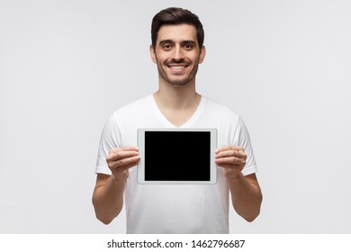 Young man holding tablet and showing blank screen with happy smile as if advising product, service or app, isolated on gray background