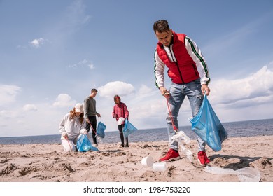 Young Man Holding Trash Bag And Collecting Rubbish On Sand Near Volunteers 