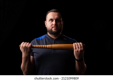 A Young Man Is Holding A Wooden Bat. Bouncer, Security Guard, Bodyguard Concept. A Large Man In A T-shirt Looks At The Camera.