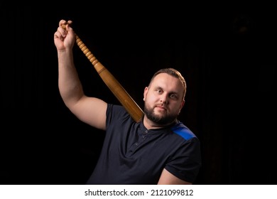 A Young Man Is Holding A Wooden Bat. Bouncer, Security Guard, Bodyguard Concept. A Large Man In A T-shirt Looks At The Camera.