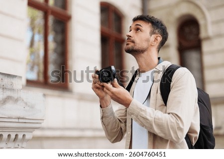 A young man holds a camera while exploring an urban setting. He appears thoughtful and curious, capturing the architectural details around him. Perfect for travel and photography themes.