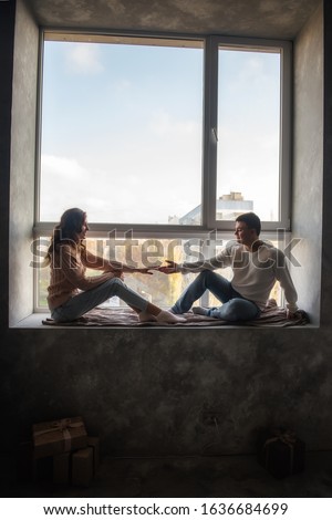 A young man holds the hand of a beautiful girl. A couple in love in warm knitted sweaters are sitting on a windowsill by a large window in their loft. Warmth and comfort, harmony in relationships.