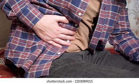 Young Man Holds His Hand To His Sore Stomach. The Second Hand Holds A Smartphone. Home Furnishings