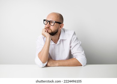 Young Man Holds His Hand Under His Chin While Sitting At A Table On A White Background.